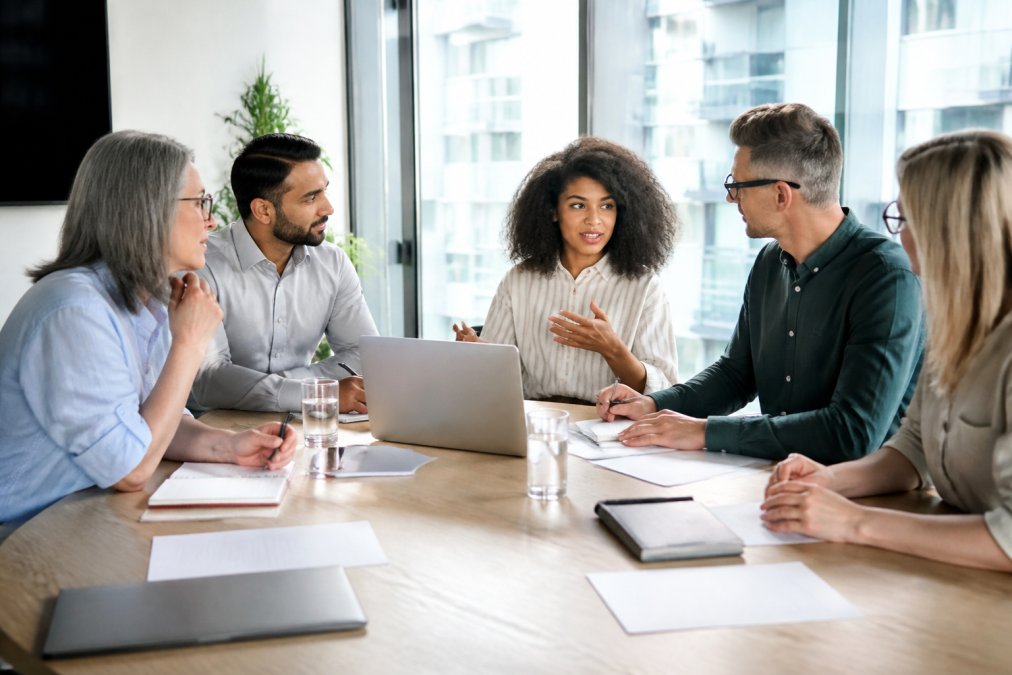 A diverse team of professionals gathered around a table in a bright office, actively discussing and collaborating with notebooks and laptops visible