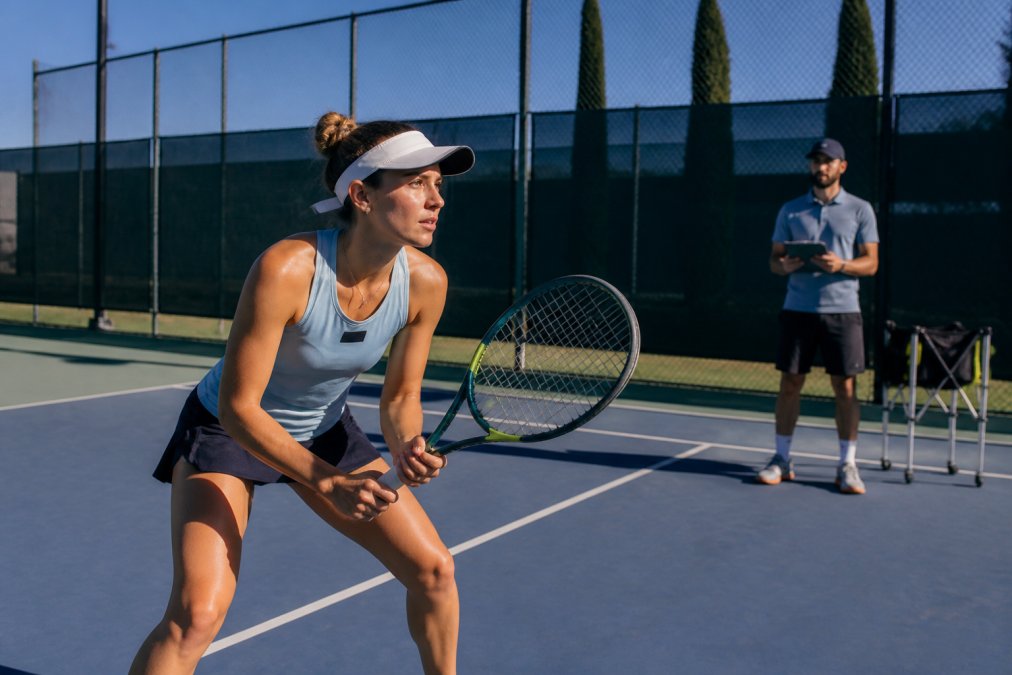 A tennis player mid-drill on a hard court, intense concentration visible on their face, a coach watching from the baseline with a clipboard, bright afternoon sunlight casting sharp shadows across the court surface