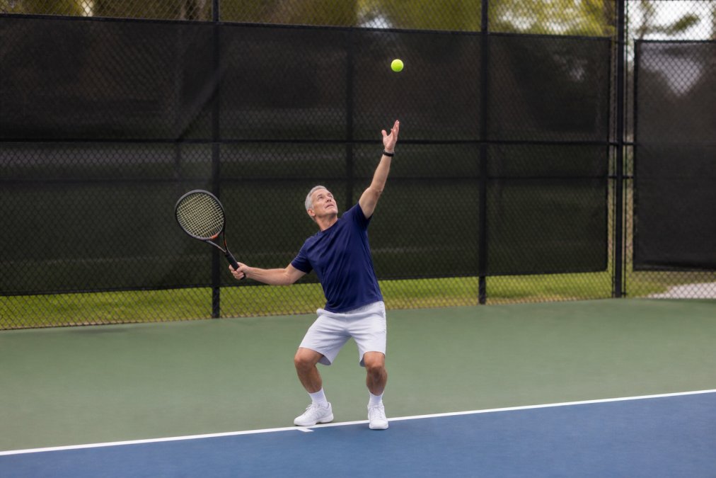 Older male tennis player in athletic stance at baseline, demonstrating loading phase with bent knees, shoulders rotated back, weight on back foot, ball toss visible at peak height above extended arm, natural outdoor court lighting