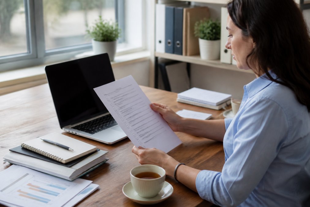 An office professional reviewing a printed process checklist at a tidy desk, with a laptop open beside them and a cup of tea nearby, in a bright, organised workspace with natural window light