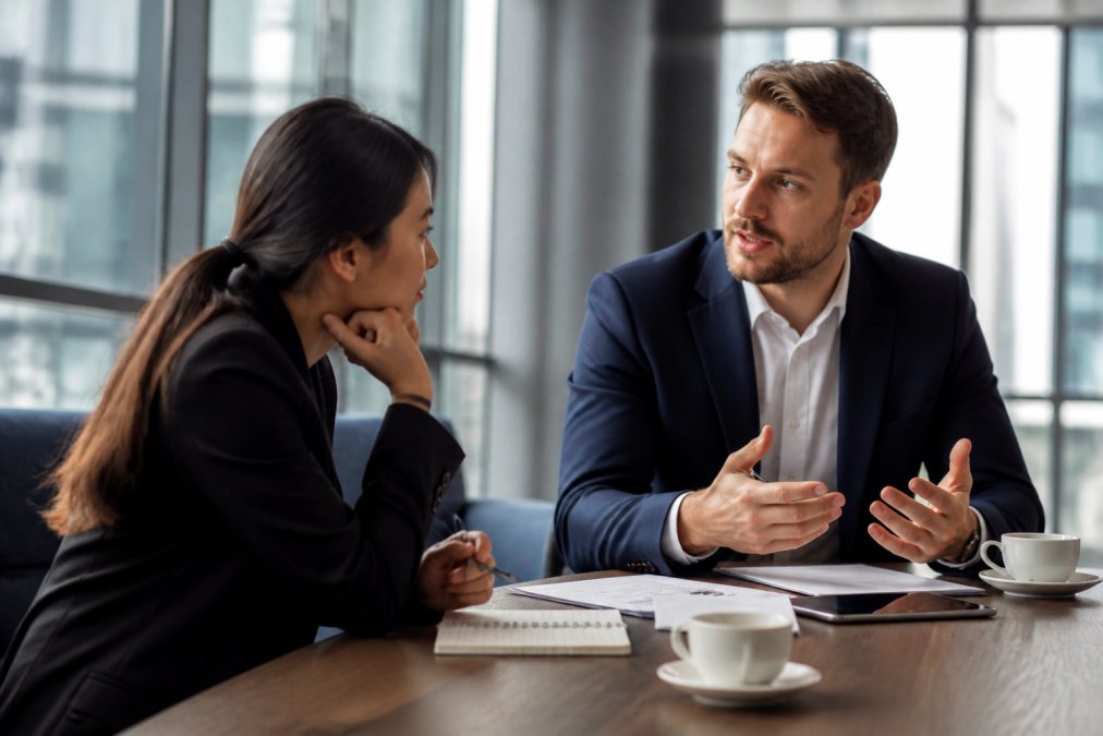 Two business professionals in a modern meeting room engaged in focused conversation, one leaning forward attentively while the other speaks, with notes and coffee visible on the table