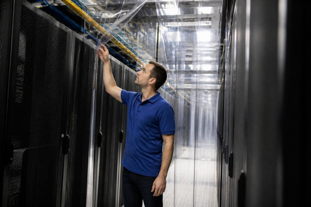 A data centre technician in a blue collared shirt inspecting server rack rows with visible hot aisle containment curtains overhead, cable management trays visible along the aisle, under bright fluorescent lighting in a live facility