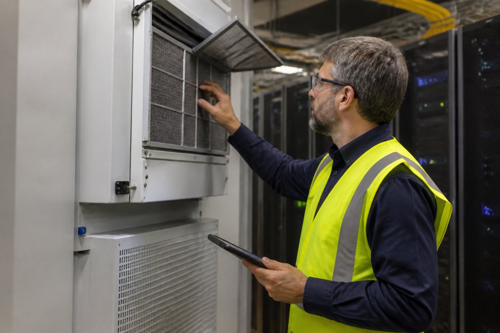 A facilities engineer wearing a high-visibility yellow vest and safety glasses performing a hands-on inspection of a precision air conditioning unit mounted in a server room, with rack enclosures visible in the background and a tablet in one hand