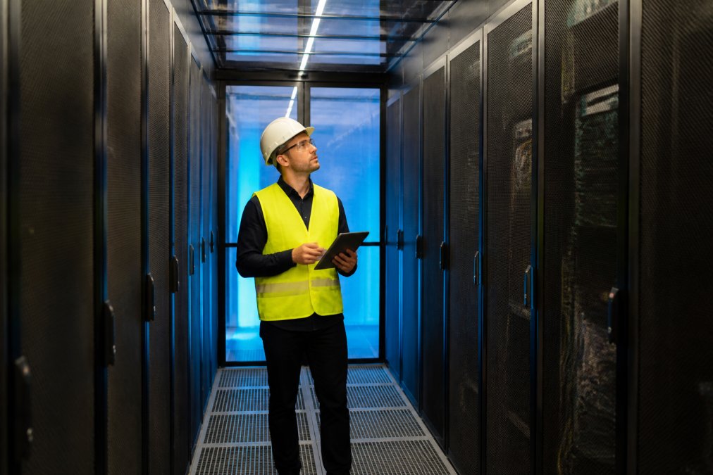 A data centre technician in a hard hat and high-visibility vest inspecting a hot aisle containment corridor between tall server racks, with bright blue cold aisle lighting visible through a transparent containment panel in the background