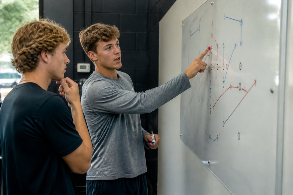 Quarterback in modern training facility studying whiteboard with defensive coverage diagrams, pointing to specific formations and coverage rotations