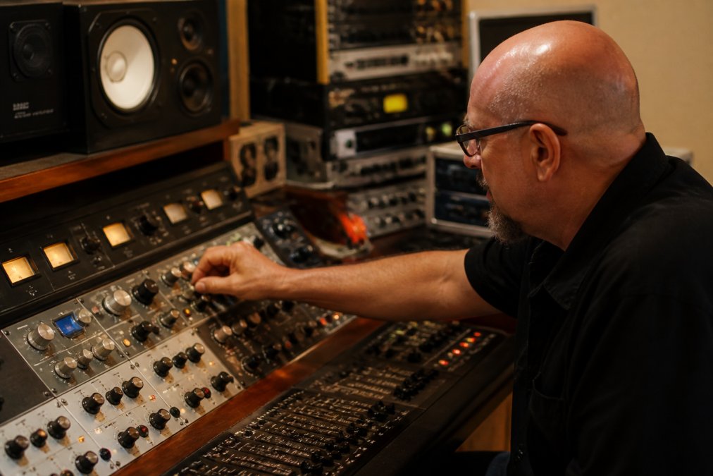 Professional audio engineer adjusting analog outboard equipment knobs on a mastering console with a DAW visible on screen in a modern studio environment