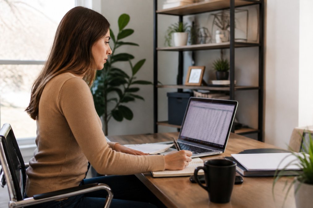 Real estate professional seated at desk in modern home office, laptop open displaying visitor contact information and notes from open house, reviewing data for follow-up strategy with natural window lighting