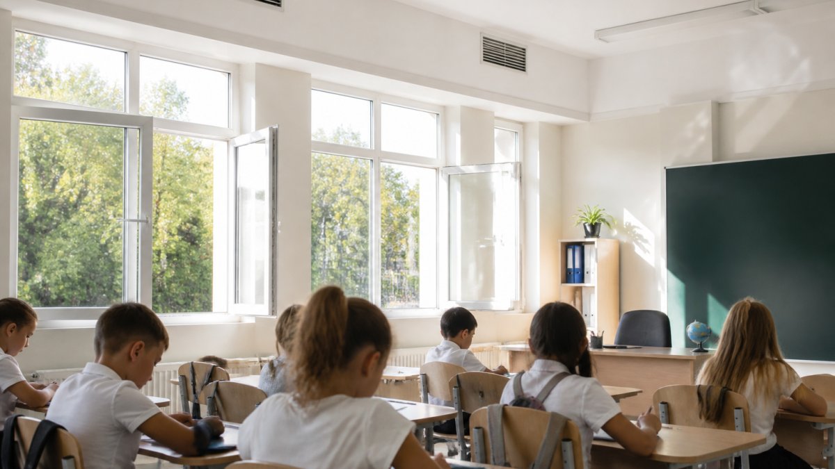 Bright school classroom with open windows, natural light streaming in, and visible air vents near the ceiling, with students at desks in a well-lit spacious environment