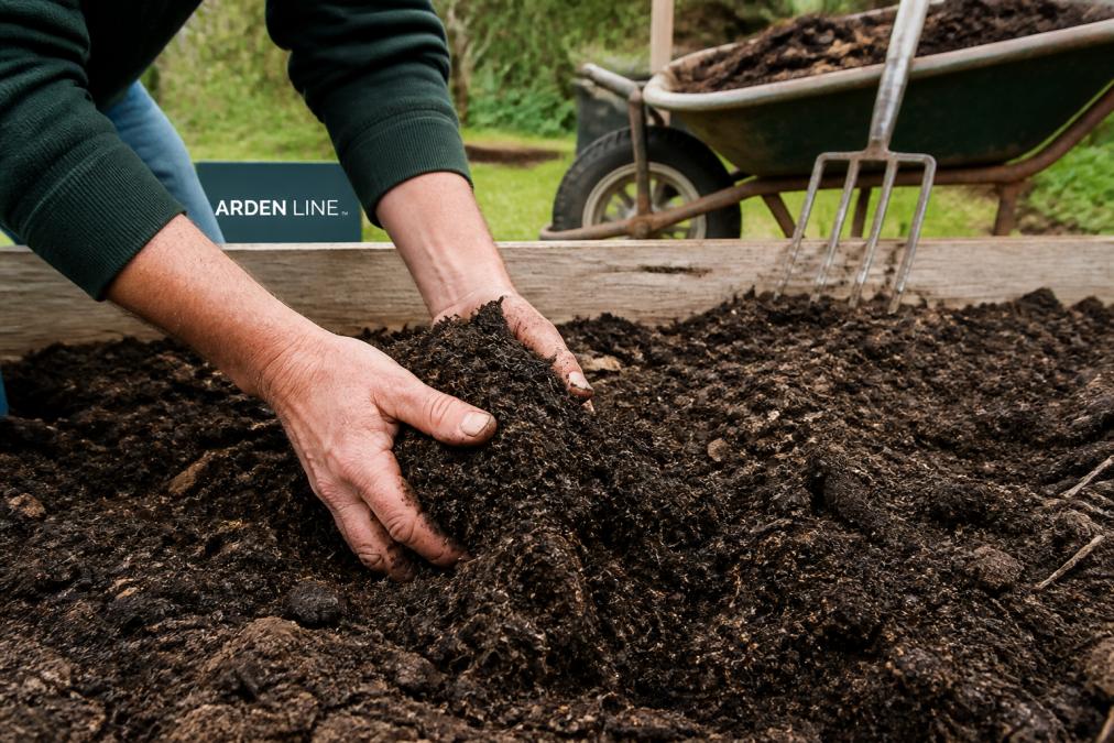 Close-up of a gardener's hands mixing dark, crumbly compost into garden soil, with a wheelbarrow and pitchfork visible in the background