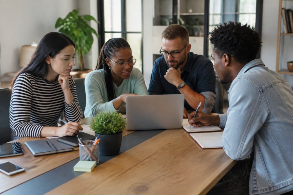 Diverse team of professionals collaborating at a modern office desk, with one person pointing at a laptop screen displaying data analytics while others take notes and discuss strategy