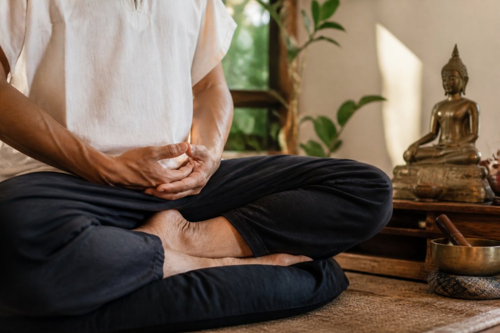 Primo piano delle mani di una persona appoggiate in grembo nel dhyana mudra, la mano destra sopra la sinistra, i pollici che si toccano leggermente, seduta su un cuscino da meditazione in una stanza scarsamente illuminata con una piccola statua buddista nelle vicinanze