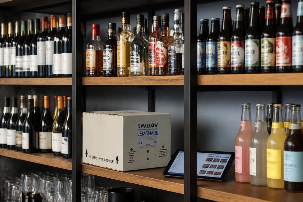 Close-up of bar shelving with organised bottles of spirits, wines, and soft drinks arranged by category, with a digital tablet displaying stock levels resting on a lower shelf in a well-lit independent bar setting