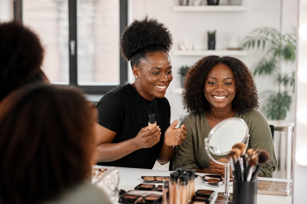 A Black woman beauty consultant demonstrating makeup products to a client at a bright, modern vanity table, both smiling and looking at a mirror reflection