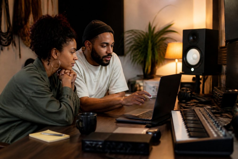 A music producer and label manager reviewing streaming analytics on a laptop in a home studio, studio monitors and a MIDI keyboard visible in the background, warm lamp lighting
