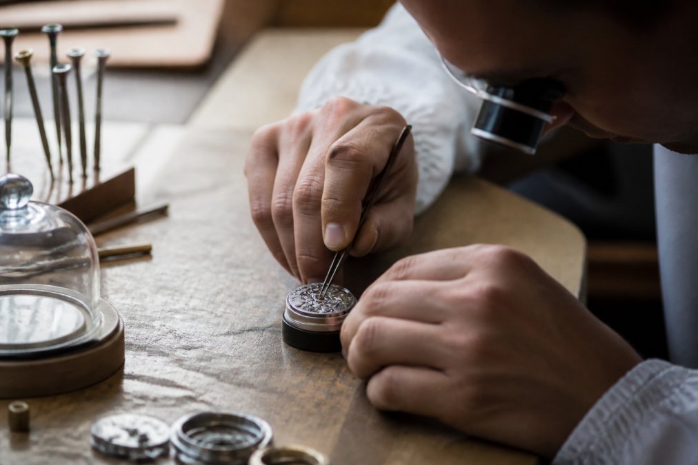 Close-up of a watchmaker's hands assembling a mechanical movement at a workbench, soft window lighting illuminating tiny gears and a loupe