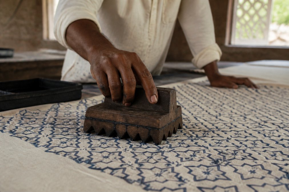Close-up of artisan's hands pressing wooden block carved with geometric patterns onto natural cotton fabric, dye dripping from block edges, traditional workshop setting with natural light