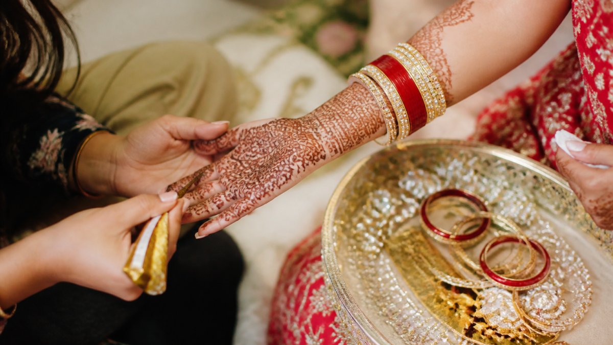 Close-up of intricate henna being applied to a bride's hands, with bangles and jewellery arranged on a decorated tray nearby in soft natural light