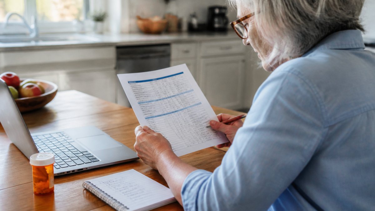 A person at a kitchen table with a laptop open to a plan formulary list, a notepad with medication names written down, and a prescription bottle nearby