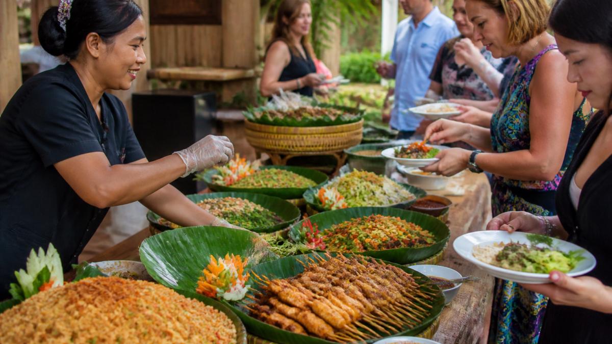 Professional caterer arranging vibrant Indonesian buffet spread with nasi goreng, sate ayam, and fresh vegetable dishes on banana leaf platters as guests serve themselves from colourful table display
