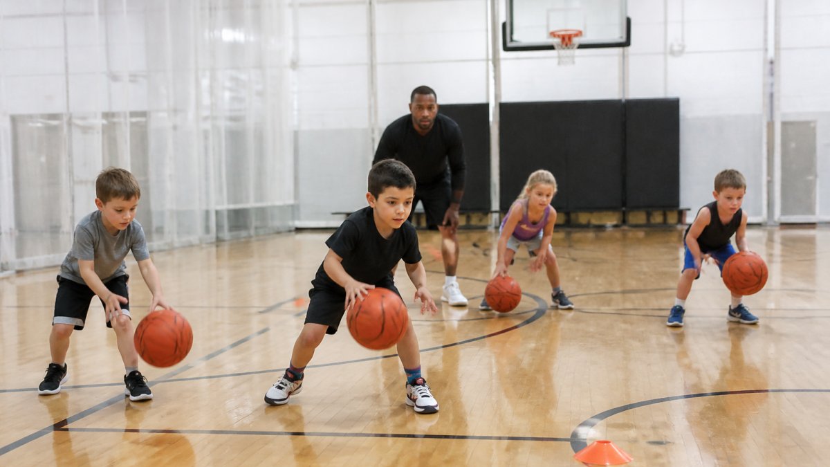 Young children aged 5-7 practicing basketball fundamentals indoors with a coach, dribbling basketballs on a full court with bright gym lighting