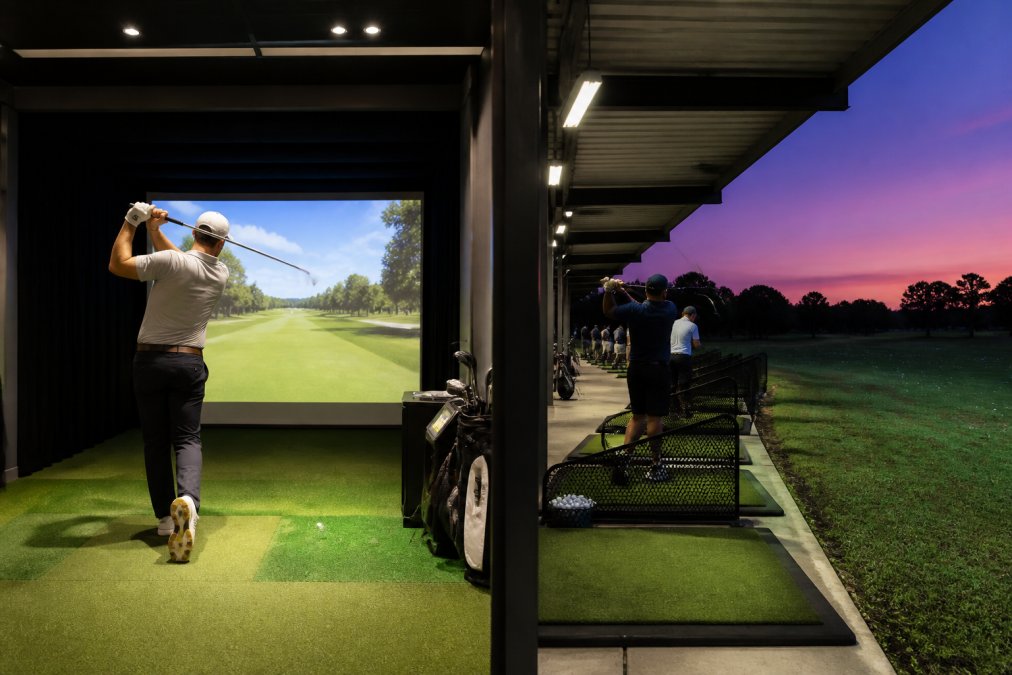A golfer mid-swing inside a private, climate-controlled simulator bay with a large projection screen showing a virtual fairway, contrasted visually with a wide outdoor driving range at dusk with several golfers hitting from covered stalls