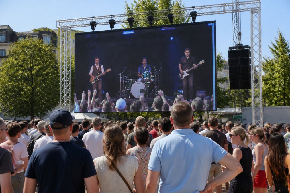 Een outdoor LED-scherm op een druk evenemententerrein overdag, met duidelijk zichtbare beelden ondanks fel zonlicht, omringd door bezoekers die naar het scherm kijken