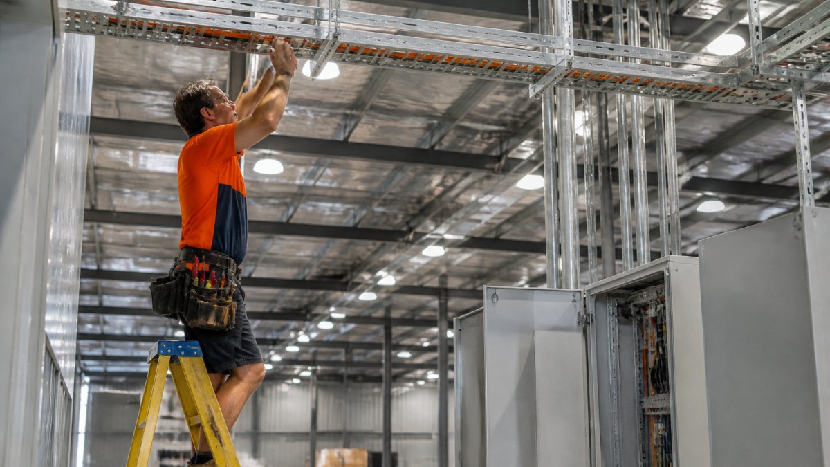 Professional electrician installing industrial cable trays and power distribution equipment in a large warehouse space, with overhead lighting fixtures and metal conduit visible in the background