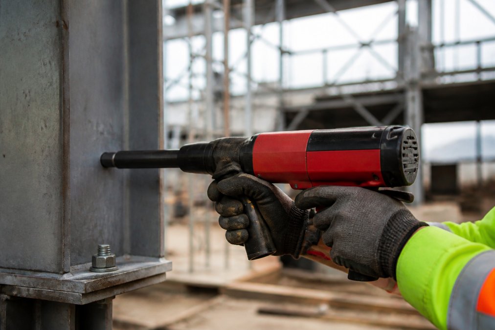 Contractor's hands operating a powder-actuated fastening tool on a structural steel beam on an active industrial jobsite, showing proper grip and positioning