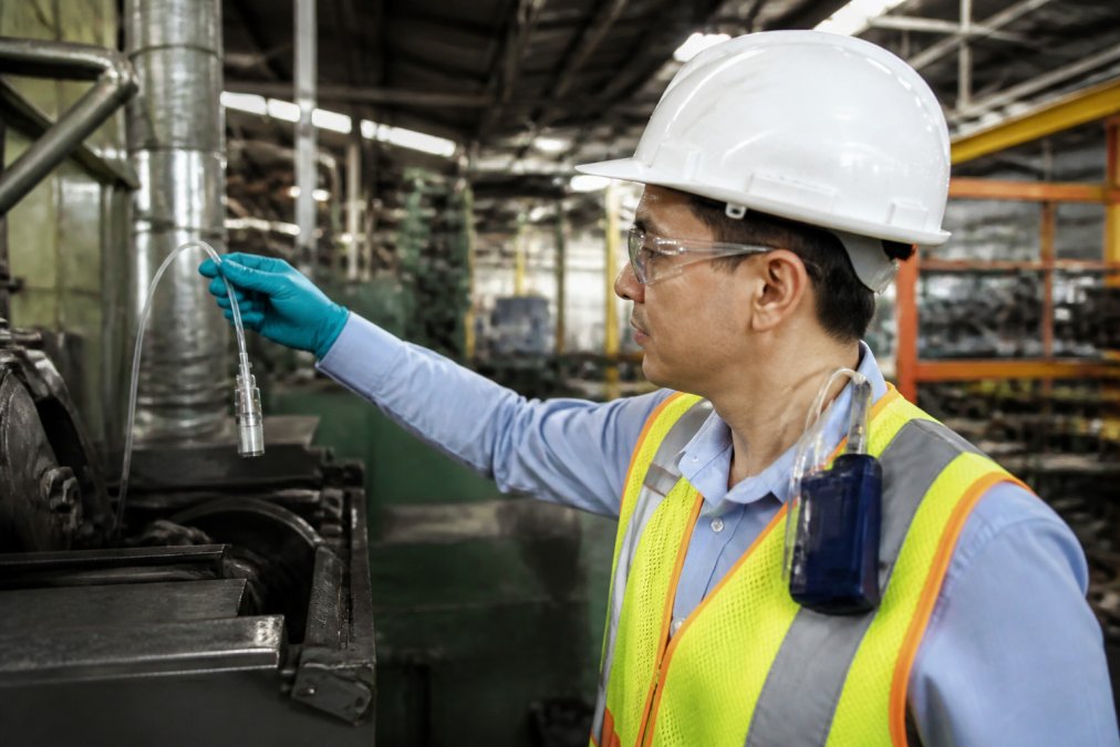 Industrial hygienist in a hard hat and safety glasses collecting an air sample near production equipment in a manufacturing facility, with sampling pump clipped to a worker's collar