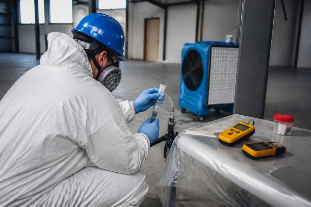 Technician collecting air quality samples and surface samples in a remediated industrial space, with HEPA filtration equipment visible in the background and moisture meters on work surfaces