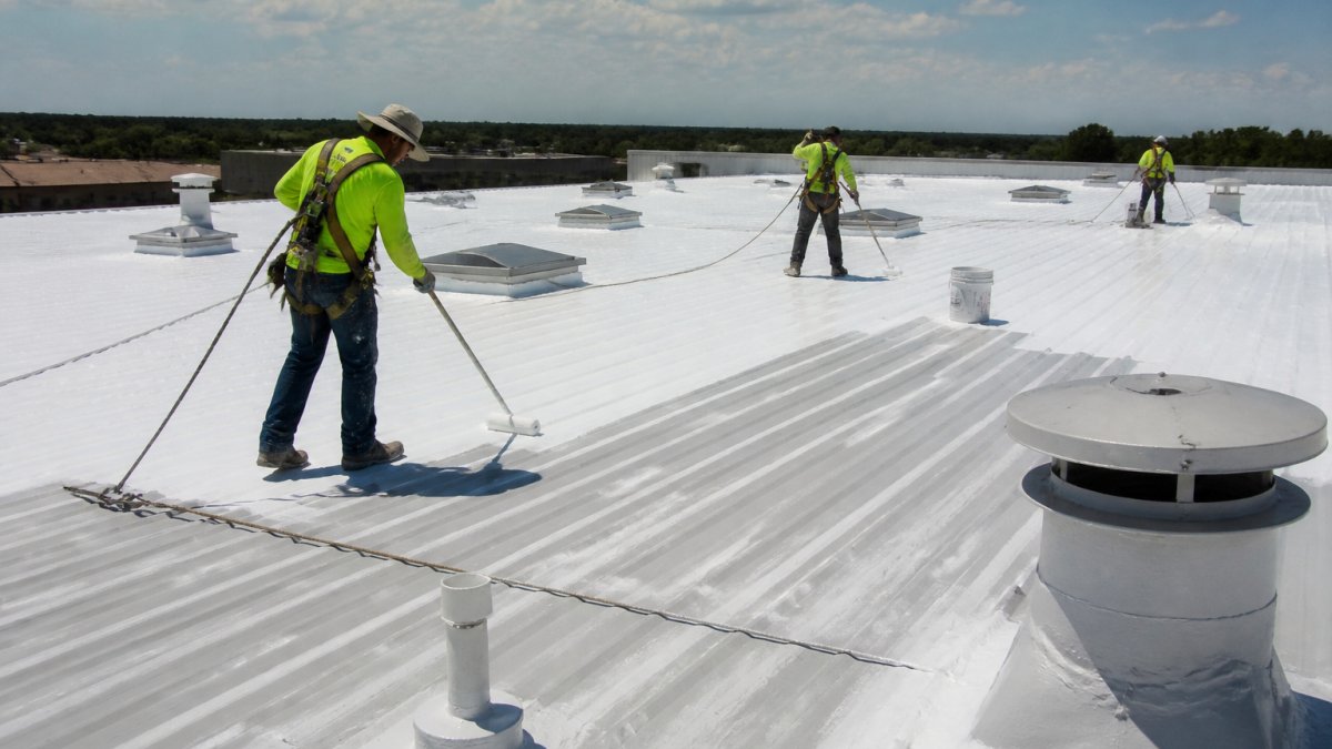 A roofing crew in safety harnesses applying a liquid silicone coating with rollers across a large warehouse metal roof, with seam details and roof penetrations visible in the foreground