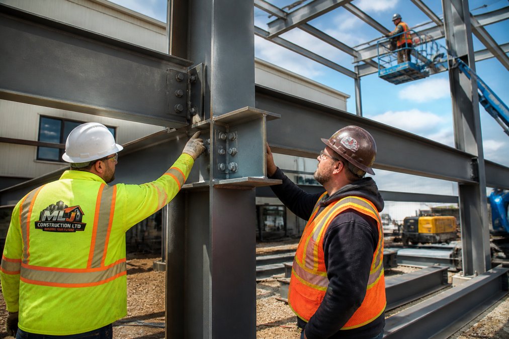 Steel frame construction site showing welded structural beams and columns with workers in safety gear and hard hats inspecting the structural connections