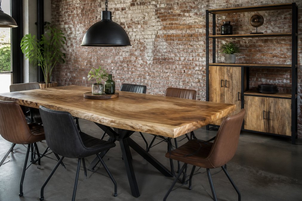 Een eetkamer met een massieve suarhouten boomstamtafel als middelpunt, omringd door stoelen met leren bekleding, een zwart stalen hanglamp erboven en een bakstenen muur op de achtergrond, warm daglicht door een hoog raam