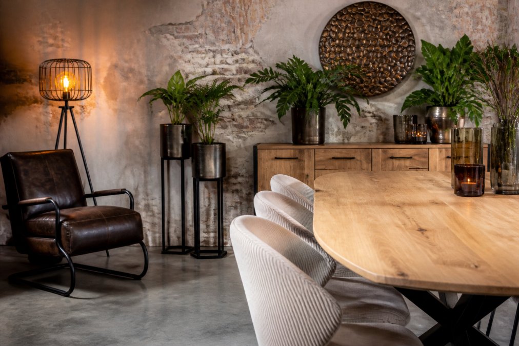 Gezellige woonkamer met een massieve mangohout eettafel, leren fauteuil in donkerbruin, stalen staande lamp met warm geel licht, houten dressoir en groene planten in metalen potten op verschillende niveaus