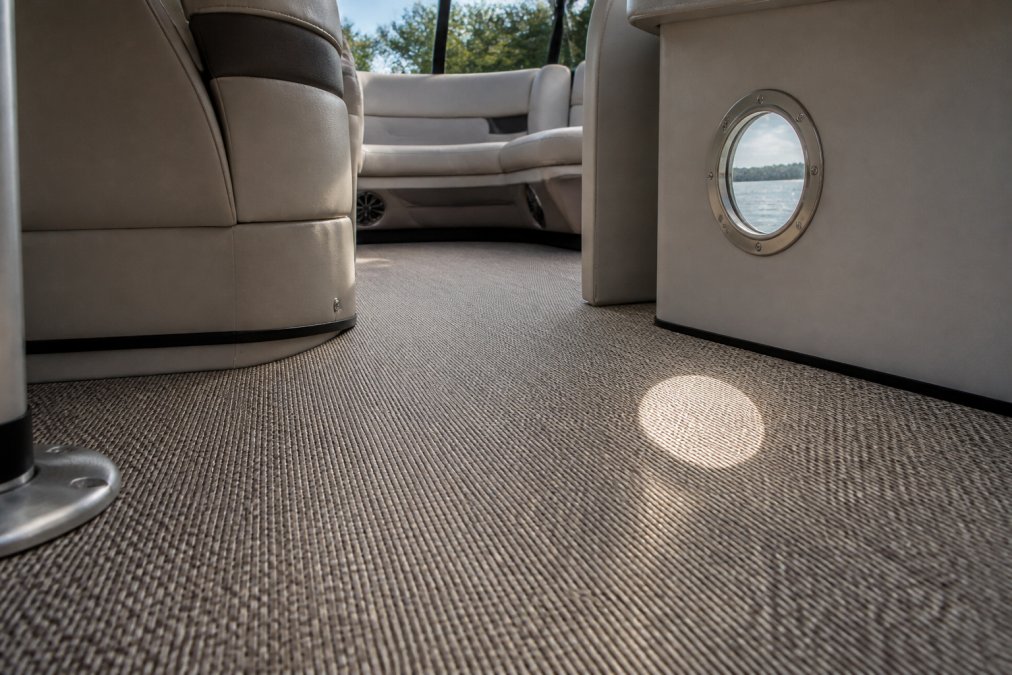 Close-up of woven vinyl flooring installed in a modern pontoon boat cabin with sunlight streaming through a porthole window, showing the texture and durability of the material in a marine environment