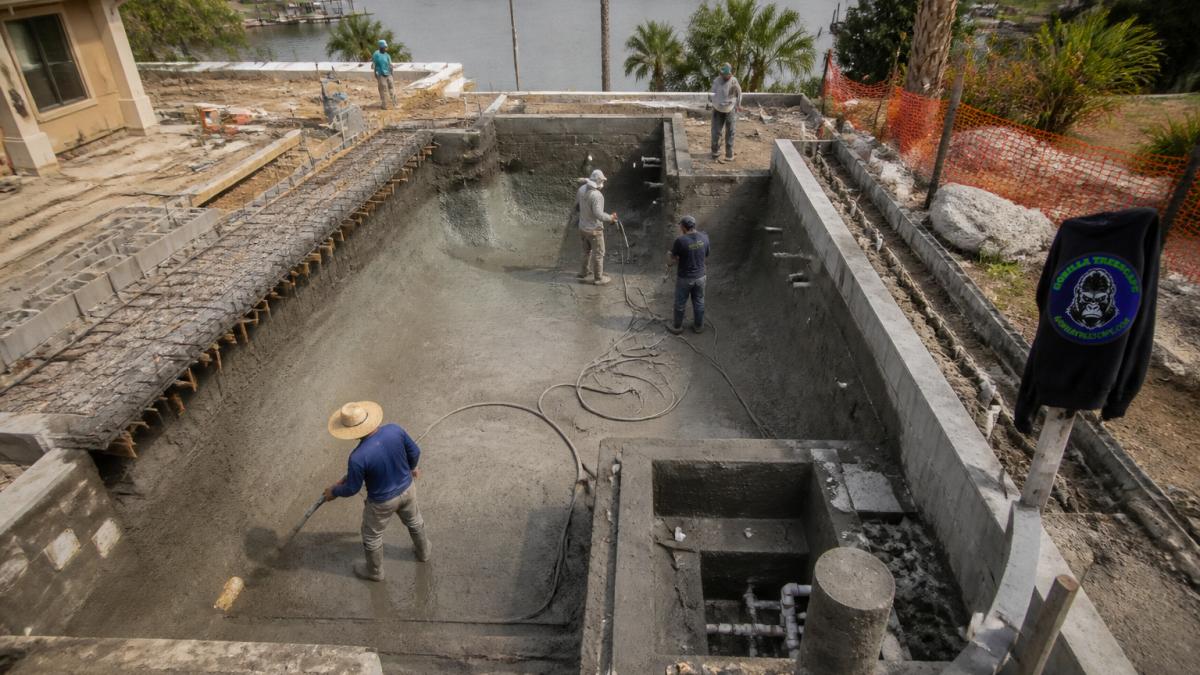 Aerial view of custom infinity pool under construction showing gunite application, catch basin infrastructure, and surrounding landscape with workers preparing the site