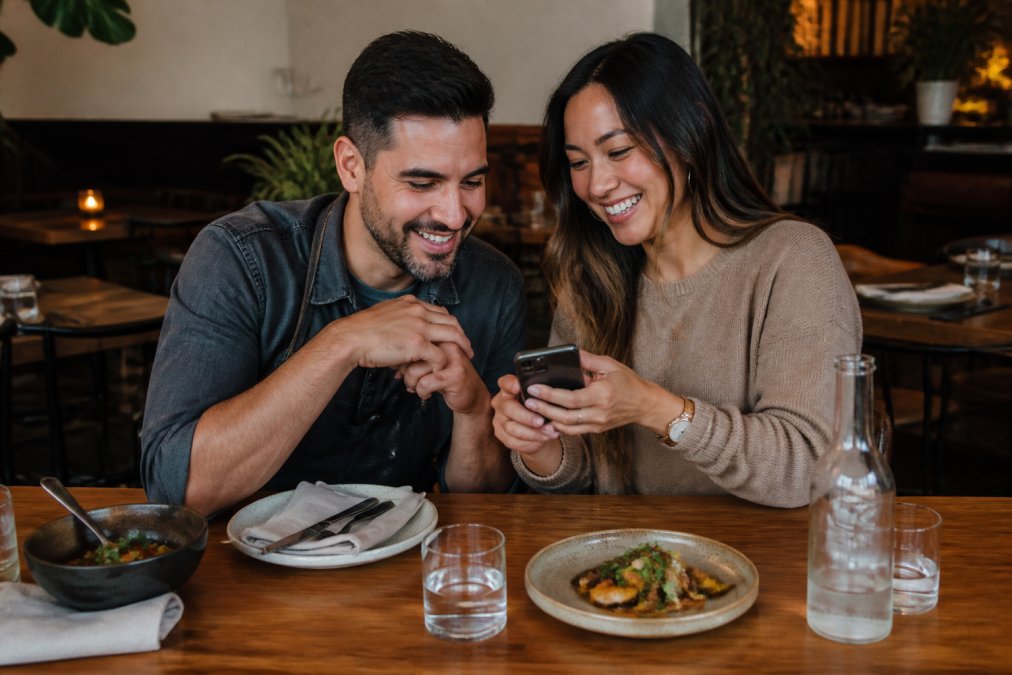 A restaurant owner and a local food content creator seated at a table reviewing a phone showing a short-form video, warm lighting, cozy dining room background