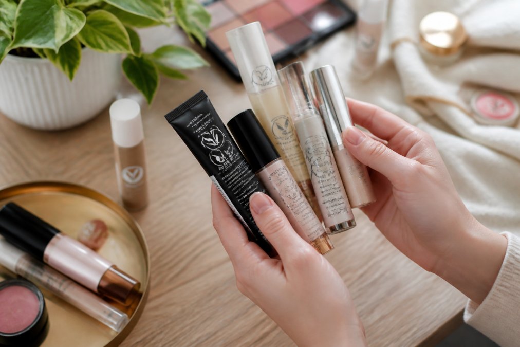 Hands holding multiple vegan makeup products with visible certification logos and labels on the packaging, arranged in natural daylight