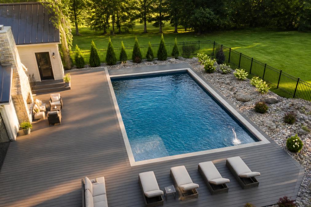 Aerial view of a completed backyard inground pool with surrounding composite decking, lounge chairs, and landscaping, showing the finished installation in afternoon sunlight