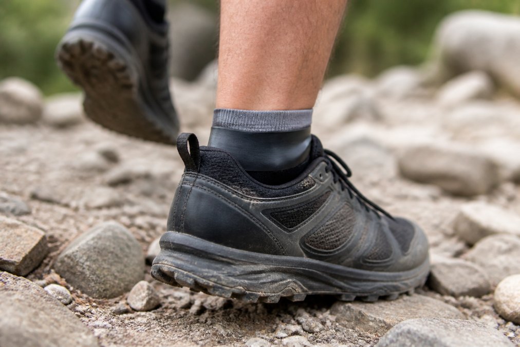Close-up of a trail runner's foot in motion on rocky terrain, showing the debris shield cuff positioned at the ankle collar with the polyurethane barrier visible and seated properly in the shoe opening
