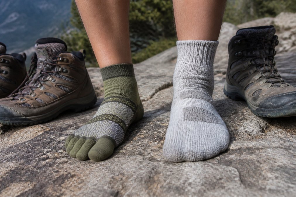 Close-up of a hiker's feet in toe socks and standard hiking socks side by side on a rocky trail, boots resting nearby, natural daylight