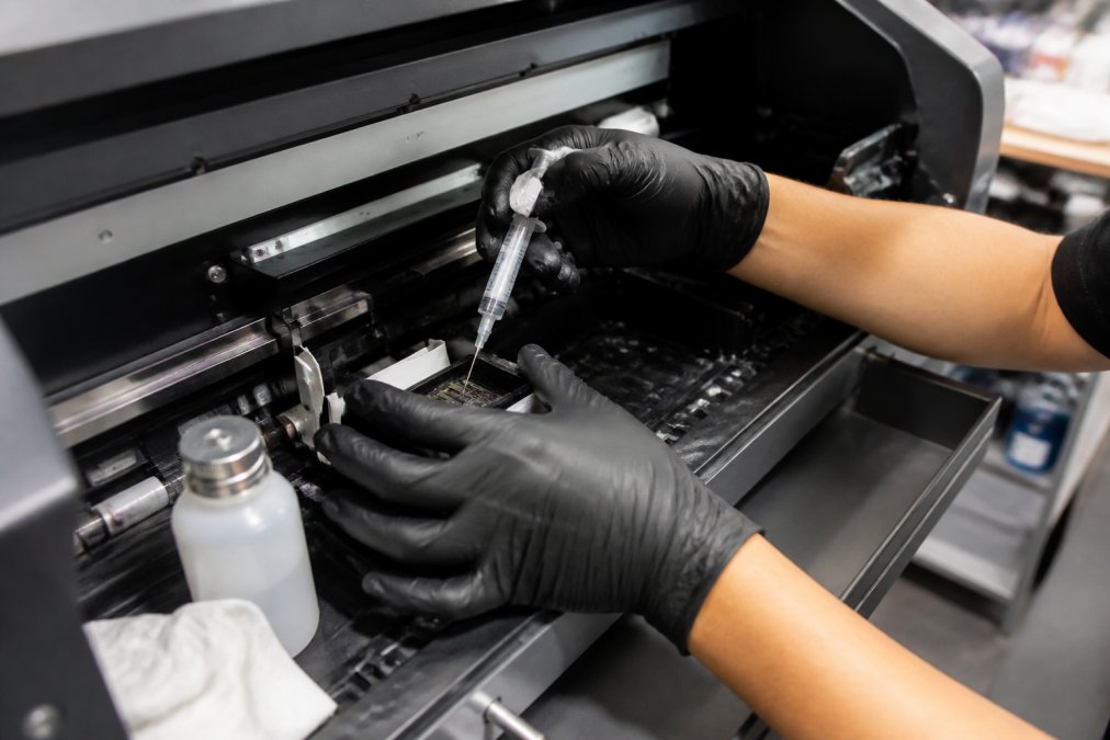 Hands performing DTF printer maintenance, cleaning print-head nozzles with precision tools and cleaning solution on an Epson wide-format printer in a production workshop