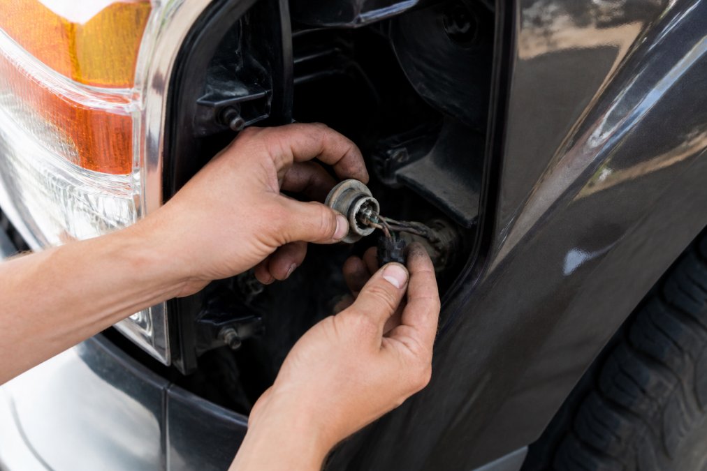 Mechanic's hands carefully disconnecting a turn signal bulb socket from a truck's front fender assembly, with the socket rotated counterclockwise and partially removed, showing the wiring harness connection