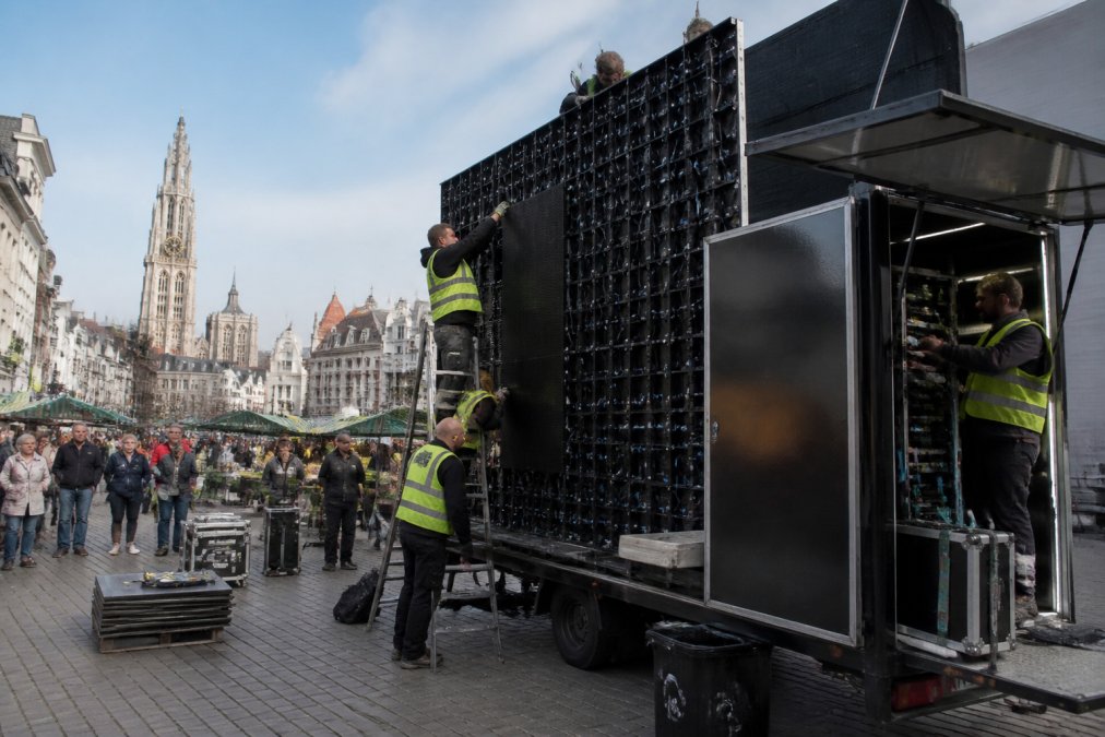 Een LED-aanhangwagen met regieruimte wordt op een stadsplein in Antwerpen opgesteld, technici in veiligheidshesjes bevestigen de panelen en regelen de bekabeling, omstanders en marktkramen op de achtergrond