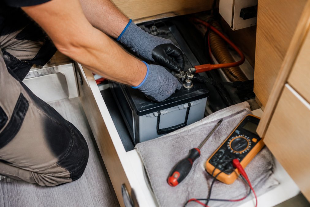 Mains gantées branchant les cosses d'une batterie auxiliaire dans un camping-car, avec outils à proximité.