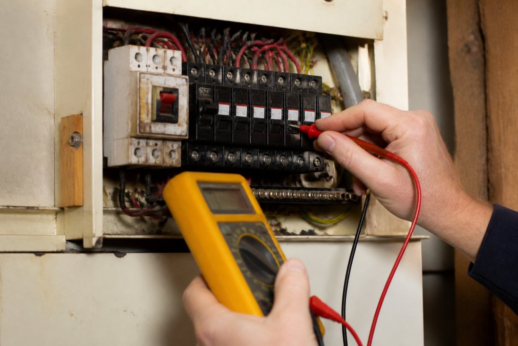 Close-up of an electrician's hands inspecting an older residential electrical consumer unit with a multimeter, examining circuit breakers and fused switches inside the panel box