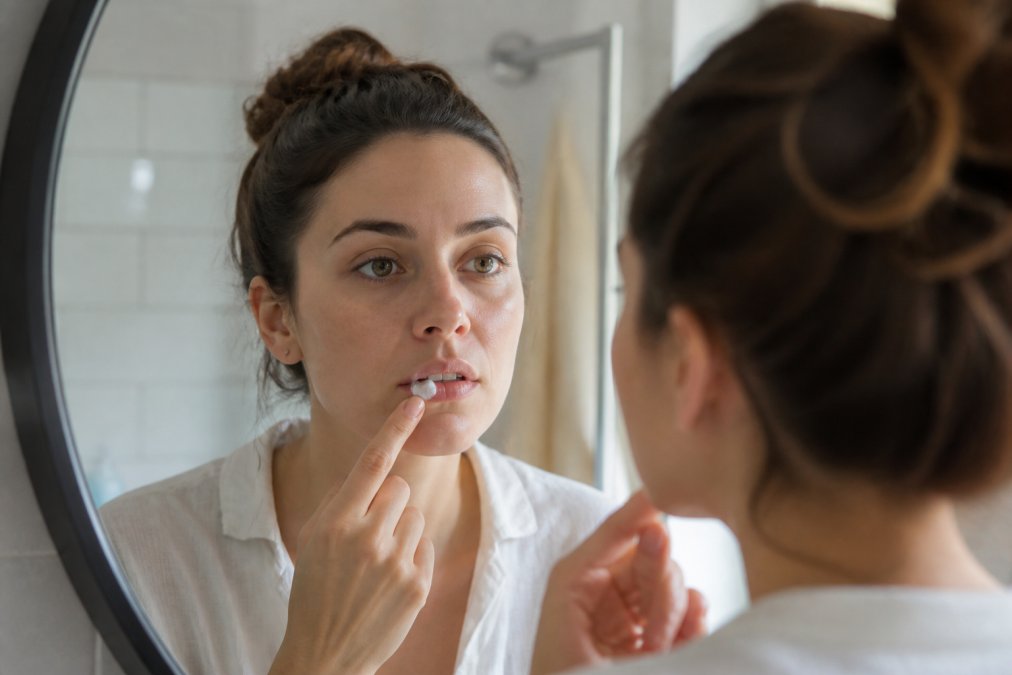 Close-up of a person applying a small amount of white topical cream to their lip with a clean fingertip, looking into a bathroom mirror with soft natural lighting