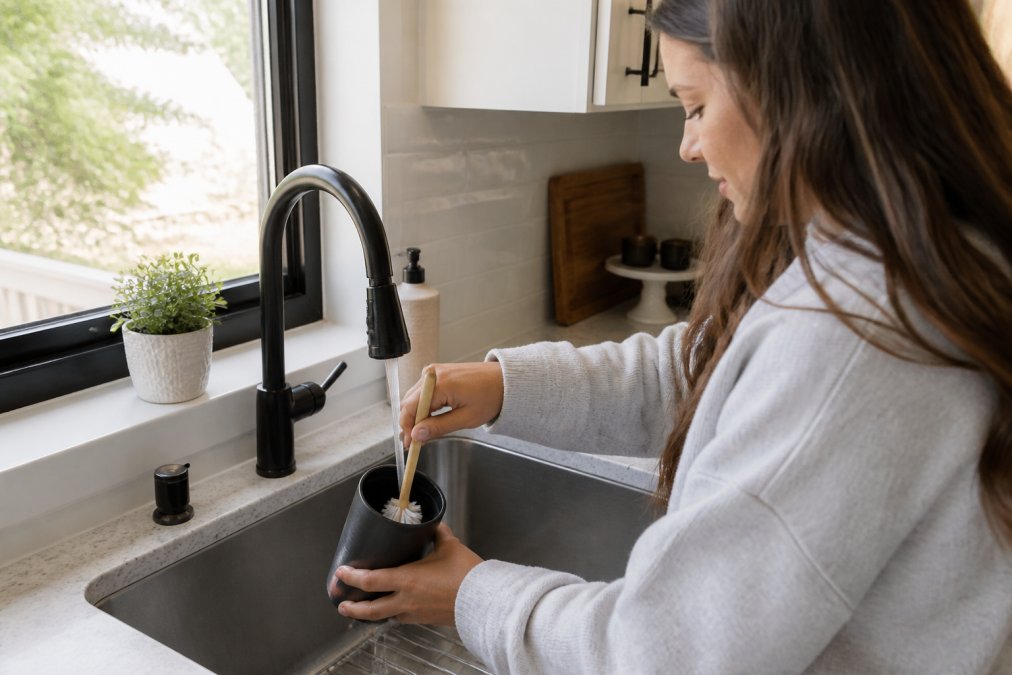 Hands washing an insulated tumbler under running water in a kitchen sink, using a soft-bristled brush to gently clean the interior surface, with natural window lighting