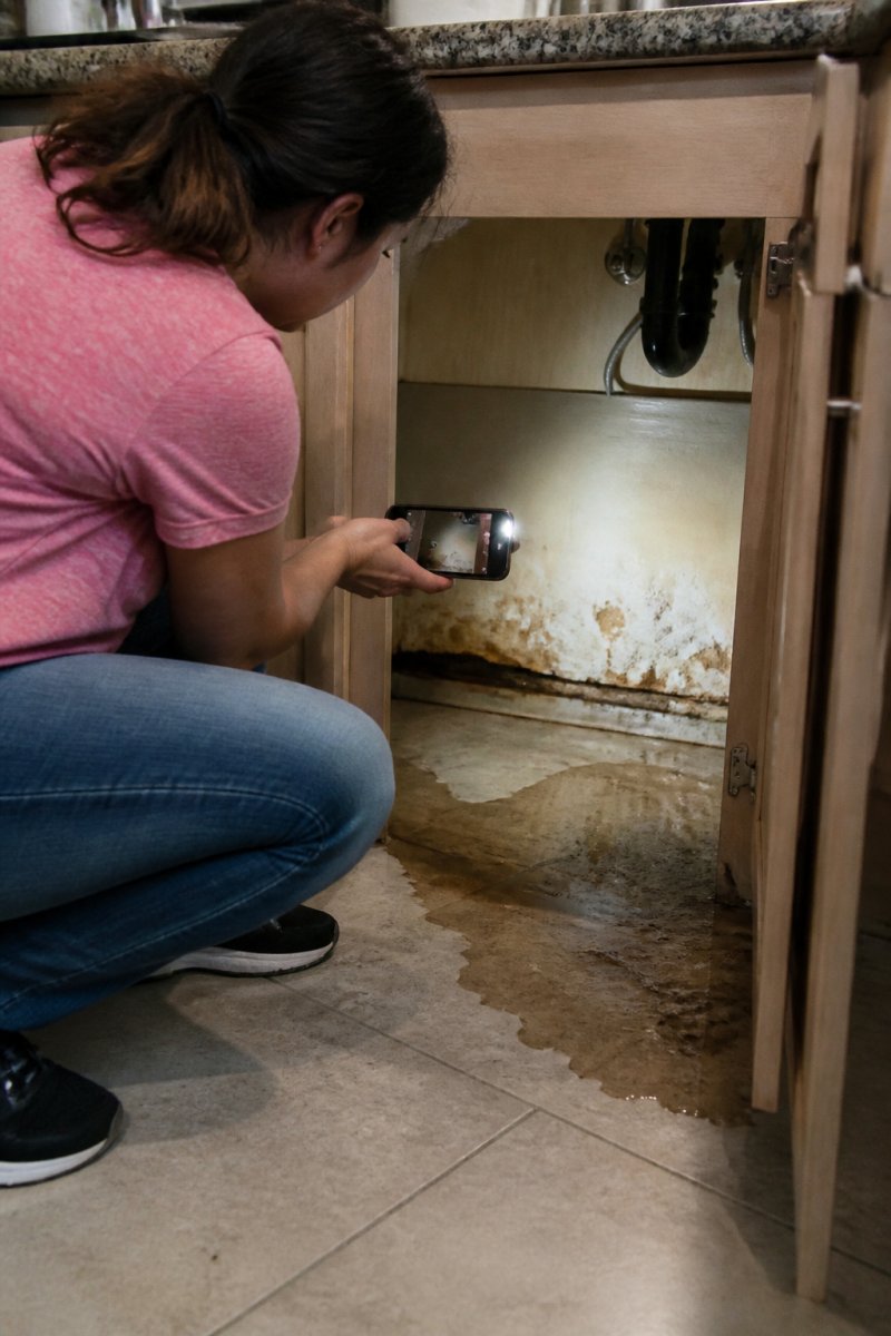 Homeowner photographing water damage under a kitchen cabinet to support a plumbing claim denial.