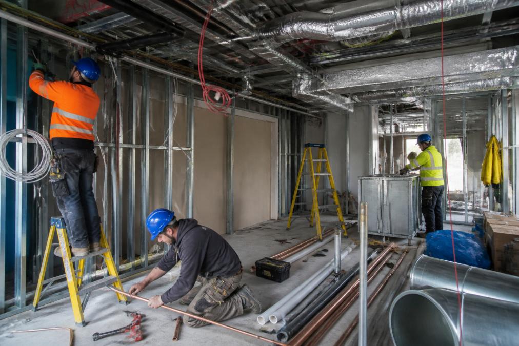 Multiple construction workers from different trades (electrician, plumber, HVAC technician) on a job site appearing to work separately without coordination, with visible scheduling conflicts and equipment in the way